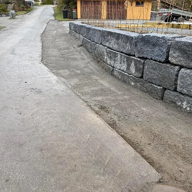 Opparbeidet skråning langs vei med støttemur av store steinblokker ved gul garasje.