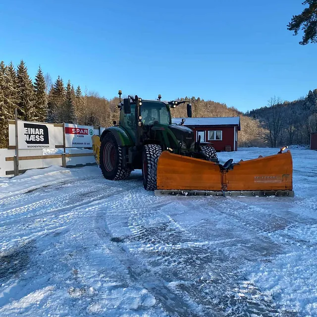 Traktor med oransje snøplog foran en rød bygning på snødekt gårdsplass.